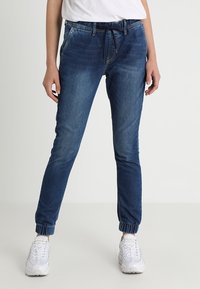 Pantalones jogger de mezclilla azul con puños elásticos y cintura ajustable con cordón. Presentan un diseño clásico de cinco bolsillos y un sutil desgaste en la tela.