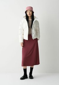 Vit pufferjacka, beige tröja, bourgognefärgad midi-kjol, svarta strumpor och svarta loafers. Modellen bär en rosa bucket hat.