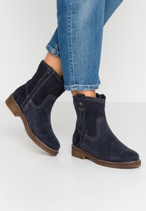 Personne portant des bottines en daim bleu foncé avec des semelles marron et un jean en denim bleu, debout sur une surface blanche.