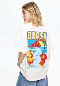 Bílé oversized tričko s barevným tropickým potiskem, na kterém je slovo "BRASIL", květy hibiskusu, hory a oceánské vlny.