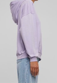 Sudadera de color púrpura claro hecha de una tela suave, con un bolsillo frontal y capucha con cordón. Ajuste holgado con puños y dobladillo de canalé.