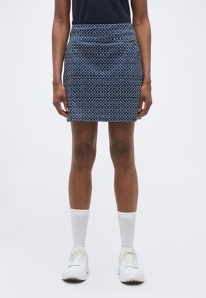 Osoba nosiaca mini sukňu s geometrickým vzorom v námorníckej modrej, bielej a čiernej farbe, s bielymi ponožkami a bielymi teniskami, stojaca vzpriamene.