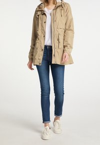 Světle béžová lehká bunda s kapucí, šňůrkami a dvěma předními kapsami, nošená přes bílou košili a modré skinny džíny.