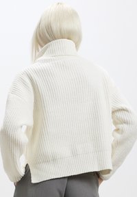 Pull à col roulé blanc, à côtes, avec une coupe décontractée. Présente un tricot épais et texturé, des épaules tombantes et une taille cropped.