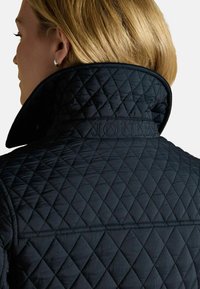 Jachetă matlasată de culoare bleumarin, cu guler înalt și branding proeminent "JOULES". Prezintă modele în romburi și o textură netedă.