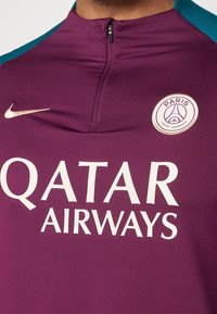 Primo piano di una maglietta sportiva bordeaux del Paris Saint-Germain con il logo bianco di Qatar Airways e il baffo Nike sul petto.