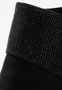 Mustat nilkkurit, joissa on pehmeä mokkapinta ja pieniä mustia paljetteja peittävä nauha, korostaen tekstuuria ja kimallusta.