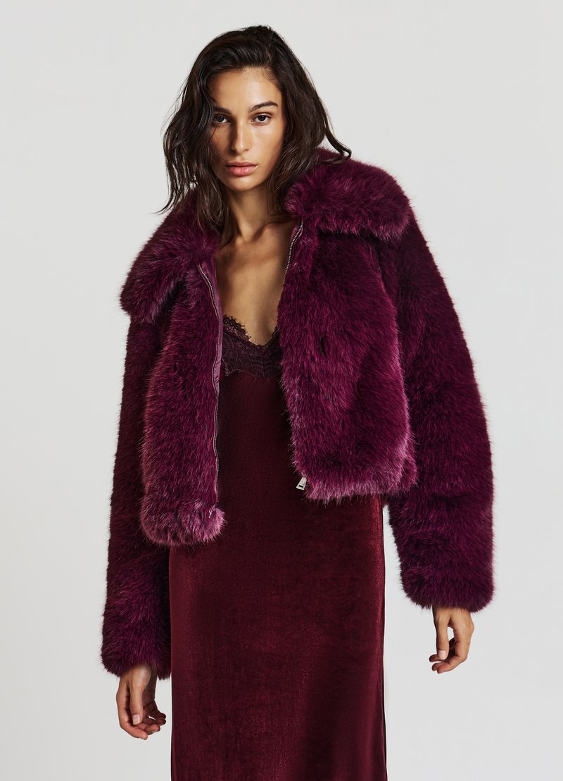 Veste en fausse fourrure, cropped, de couleur magenta profond avec une texture pelucheuse et un col surdimensionné, assortie à une robe en soie bordeaux. Détail de fermeture éclair argenté.