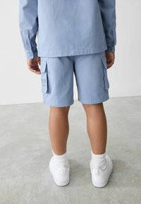 Ljusblå cargoshorts med sidofickor och en tryckt logotyp. Kombinationen matchas med vita sneakers och ankelsockor, sedd bakifrån.