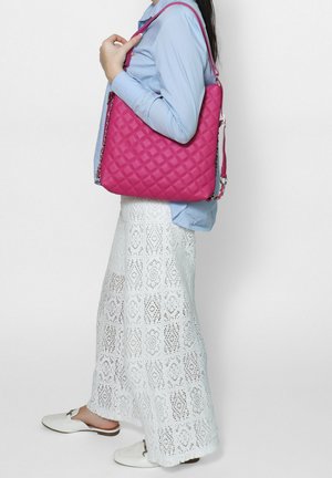 Femme portant un pantalon en dentelle blanche et une chemise bleue, tenant un grand sac à main matelassé rose sur l'épaule.