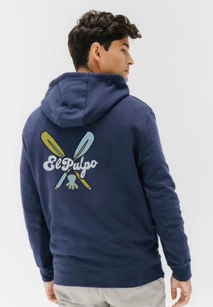 Hombre vestido con sudadera con capucha azul marino con el texto "El Pulpo" y un gráfico de remos amarillos y azules cruzados en la espalda, mirando de lado.