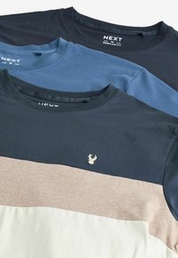 Tre T-shirts stablet, med en navy top, en blå mellem, og en beige og hvid bund, med et lille broderet logo på forsiden.