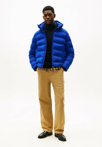 Blå pufferjakke med hætte, quiltet design, iført over en sort sweater. Sammen med beige bukser og sorte sko.