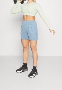 Top corto de manga larga verde claro, pantalones cortos azules con cinturilla elástica, zapatillas deportivas negras con detalles en blanco, pesas en las manos.