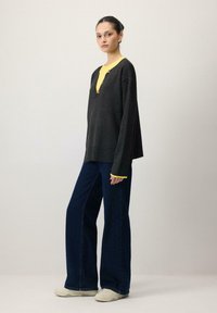 Pull oversize gris foncé avec une couche intérieure jaune, doté d'un col en V et de boutons. Associé à un jean bleu foncé à jambes larges et des baskets blanches.