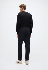 Μαύρο φούτερ με στρογγυλή λαιμόκοψη συνδυασμένο με σκοτεινό παντελόνι. Το ντύσιμο έχει άνετη εφαρμογή. Κρεμ αθλητικά παπούτσια με σκούρες λεπτομέρειες ολοκληρώνουν την εμφάνιση.