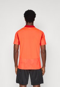 Rdeča športna majica s kratkimi rokavi, strukturo na hrbtni strani in črnim ovratnikom. Kombinirana z črnimi kratkimi hlačami. Brez vidnih logotipov ali vzorcev.