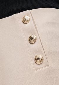 Μπεζ ένδυμα με τρία διακοσμητικά χρυσά κουμπιά τοποθετημένα κάθετα πάνω σε υφή ύφασμα.