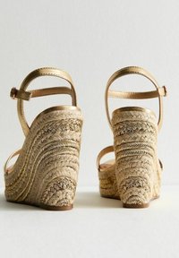 Sandali a cuneo con cinturini metallici dorati, tacco in materiali naturali intrecciati, texture curva e fibbia alla caviglia per una vestibilità sicura.