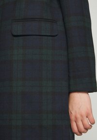 Kabát z vlněné směsi s tmavě modrým a zeleným kostkovaným vzorem. Design zahrnuje kapsu s klopou a strukturovaný detail na rameni.
