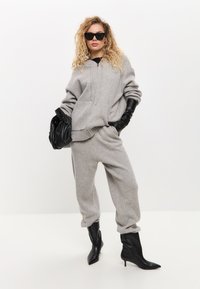 Grijze sweatshirt en joggingsbroek, gebreide stof met ribbels aan de mouwen, gecombineerd met zwarte handschoenen en spitse enkellaarsjes. Zwarte zonnebril en handtas.