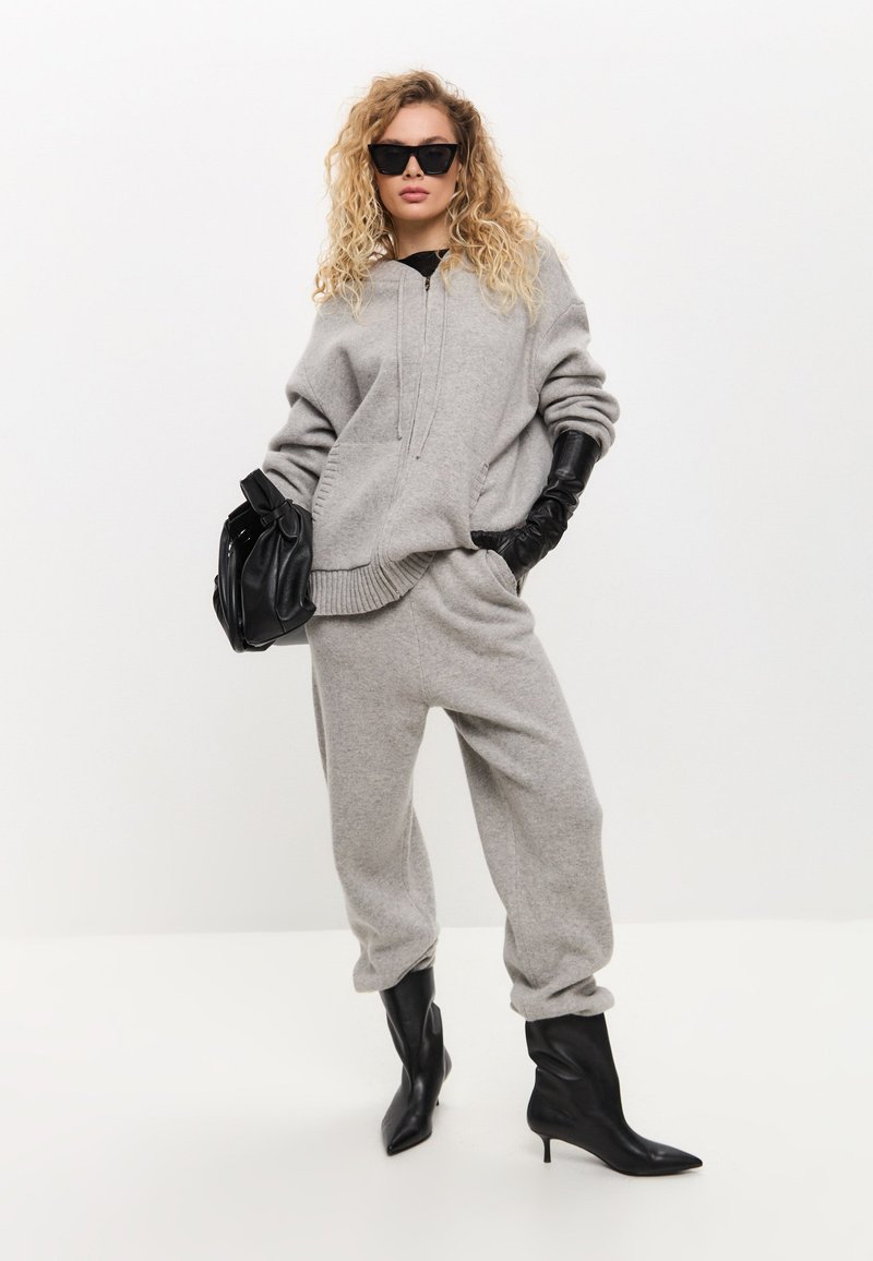Grijze sweatshirt en joggingsbroek, gebreide stof met ribbels aan de mouwen, gecombineerd met zwarte handschoenen en spitse enkellaarsjes. Zwarte zonnebril en handtas.