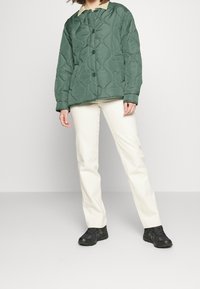 Giacca trapuntata verde con colletto color crema, abbinata a pantaloni bianchi e scarpe da ginnastica nere. Design semplice e dalla vestibilità rilassata.
