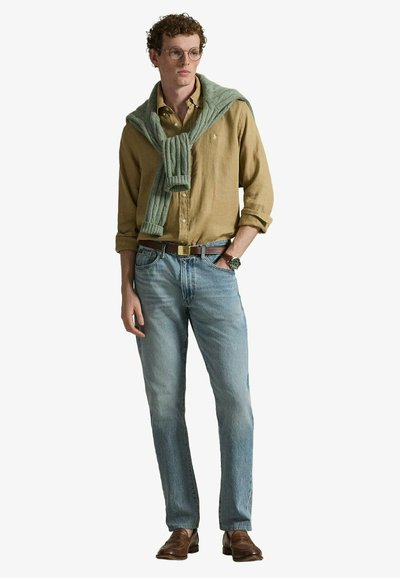 Homme aux cheveux bouclés et lunettes porte une chemise moutarde, un pull vert posé sur les épaules, un jean bleu, une ceinture marron, une montre et des chaussures marron.