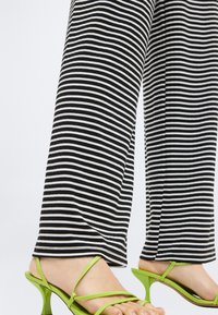 Ben iført sort-hvide bukser med horisontale striber og neongrønne højhælede sandaler med tynde stropper.