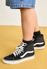 Baskets montantes noires avec des accents blancs, tissu texturé et lacets blancs. Portées avec des chaussettes noires montant à la cheville et un short en denim, sur un fond jaune.