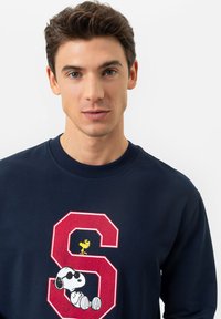 Jeune homme aux cheveux marron courts portant un sweat-shirt navy arborant un grand "S" rouge avec les personnages Snoopy et Woodstock.