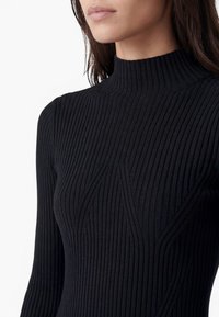 Frau trägt einen eng anliegenden schwarzen gerippten Pullover mit hohem Kragen und langen Ärmeln vor einem schlichten weißen Hintergrund.