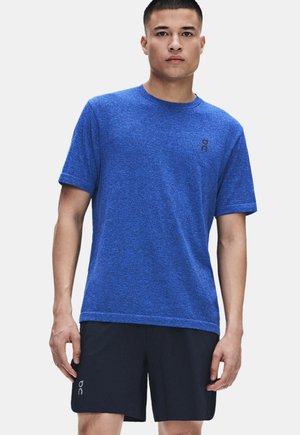 Mann in blauem kurzärmeligem Hemd und schwarzen Shorts mit kleinen Logos, steht vor einem einfarbigen hellen Hintergrund und schaut nach vorne.