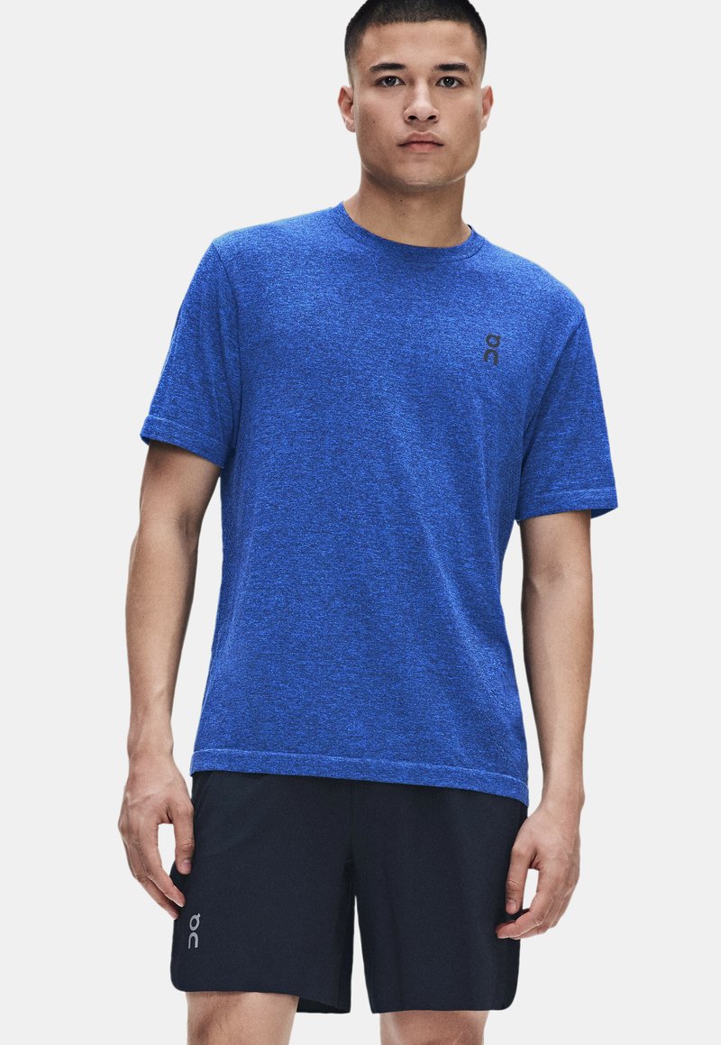 Homme debout, portant une chemise bleue à manches courtes et un short noir avec de petits logos, faisant face à un fond clair uni.