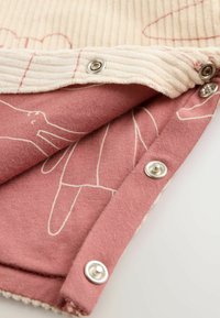 Close-up van beige ribfluwelen stof met drukknoopjes overgevouwen op stof in stofroze met witte lijntekeningen.