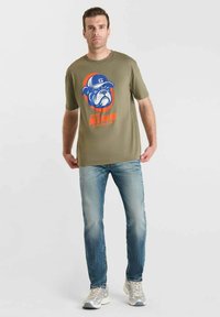 Hombre con una camiseta verde oliva con un gráfico de bulldog y texto, jeans de mezclilla claros y zapatillas blancas, de pie frente a un fondo liso.