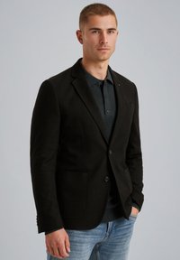 Zwarte op maat gemaakte blazer met een gestructureerde afwerking, twee voorzakken en een revers, gedragen over een donkere polo en lichte denim.