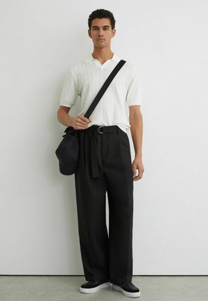 Noor mees, kes kannab valget tekstuuriga lühikeste varrukatega polot, musti laia jalaga pükse vöögaga, musta risti üle keha kantavat kotti ja musti tossusid valgete talladega.