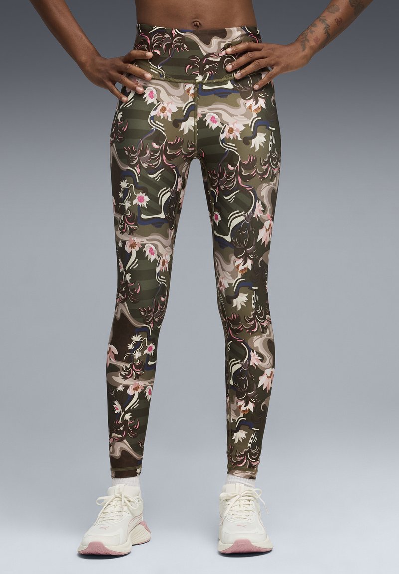 Rankos ant liemens, dėvint aukšto liemens gėlėtus, kamufliažo raštą turinčius leginsus, derinamus su baltais sportbačiais abuoti pilko fono.