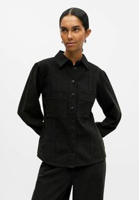Camicia in denim nero con colletto, chiusura con bottoni, due tasche sul petto e maniche lunghe. Presenta un design strutturato e dettagli di cucitura.