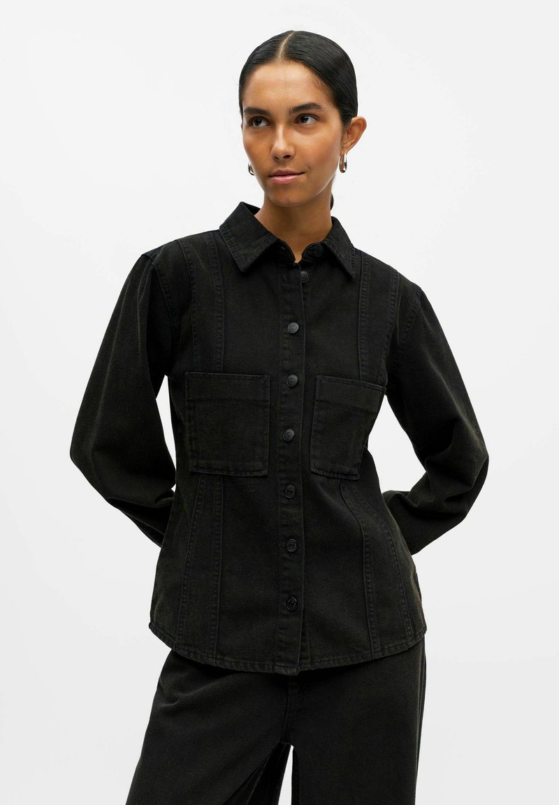 Chemise en denim noir avec un col, fermeture à boutons sur le devant, deux poches poitrine et manches longues. Présente un design structuré et des détails de couture.