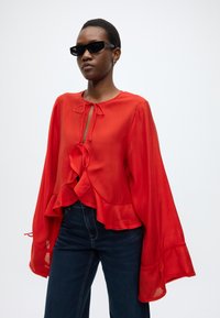 Κόκκινη διάφανη μπλούζα με μακριά μανίκια με βολάν, κλείσιμο με δέσιμο στο λαιμό και στρωτό τελείωμα. Συνδυασμένη με σκούρο τζιν. Μινιμαλιστικός σχεδιασμός.