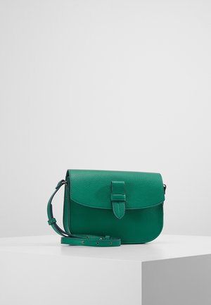 Groene leren crossbodytas met verstelbare riem en voorflap met gestikte gespdetails, tentoongesteld op een wit voetstuk tegen een effen achtergrond.