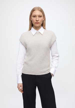 Jeune femme aux cheveux blonds raides portant une chemise blanche à col sous un gilet sans manches en tricot beige clair et un pantalon noir.
