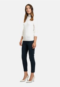 Weißer Rollkragenpullover mit langen Ärmeln, kombiniert mit dunkelblauen Hosen und weißen High Heels. Die Form ist figurbetont und betont die Silhouette.