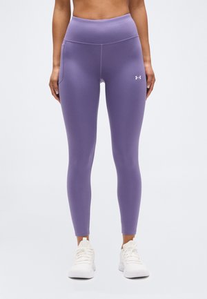 Vrouw die paarskleurige leggings met hoge taille en zijzakken draagt, gecombineerd met witte sport schoenen, staand tegen een effen lichte achtergrond.