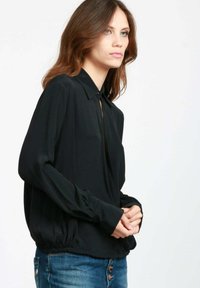 Blusa nera con colletto, maniche lunghe e vestibilità ampia. Tessuto morbido con una leggera lucentezza. Indossata con jeans in denim con bottoni.