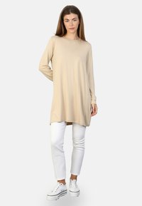 Tunique beige à manches longues avec un col rond, texture douce, fentes latérales à l'ourlet, associée à un pantalon blanc et des sneakers blanches.