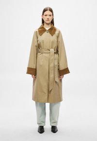 Beež värvusega trentšmantel pruunide kordurookraede ja mansettidega, vöökohal rihm, kantud hele siniste pükste ja mustade kingadega.