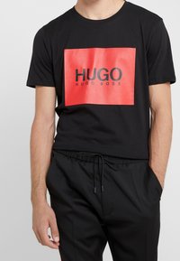 Černé bavlněné tričko s červeným obdélníkovým logem uprostřed, na kterém je text "HUGO" a pod ním "HUGO BOSS" v černé barvě. Krátké rukávy.